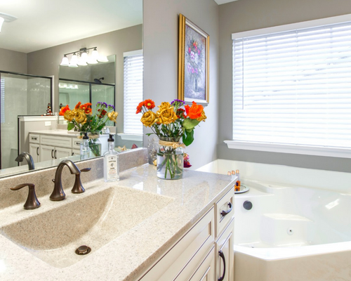 Bright bathroom with vanity, large mirror, flowers, and a sunlit bathtub window.