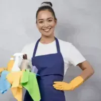 Smiling cleaner in blue apron and yellow gloves holding cleaning supplies with colorful cloths