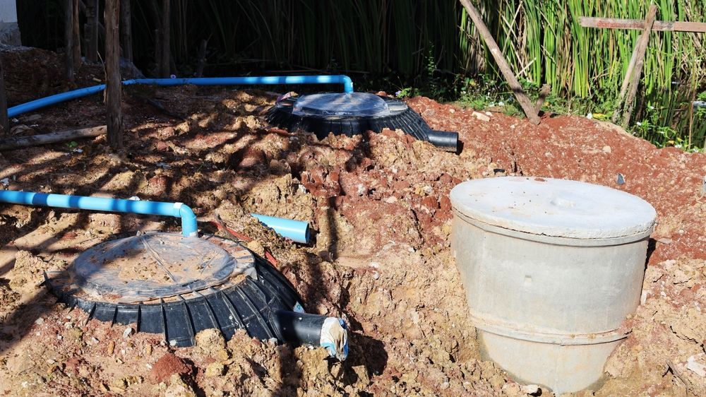 Large white bucket beside black pipe fittings and blue hoses in a dirt excavation area
