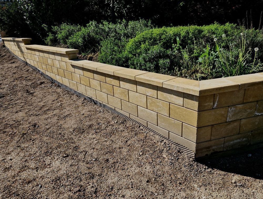Tan brick retaining wall around a raised shrub bed beside a dirt yard