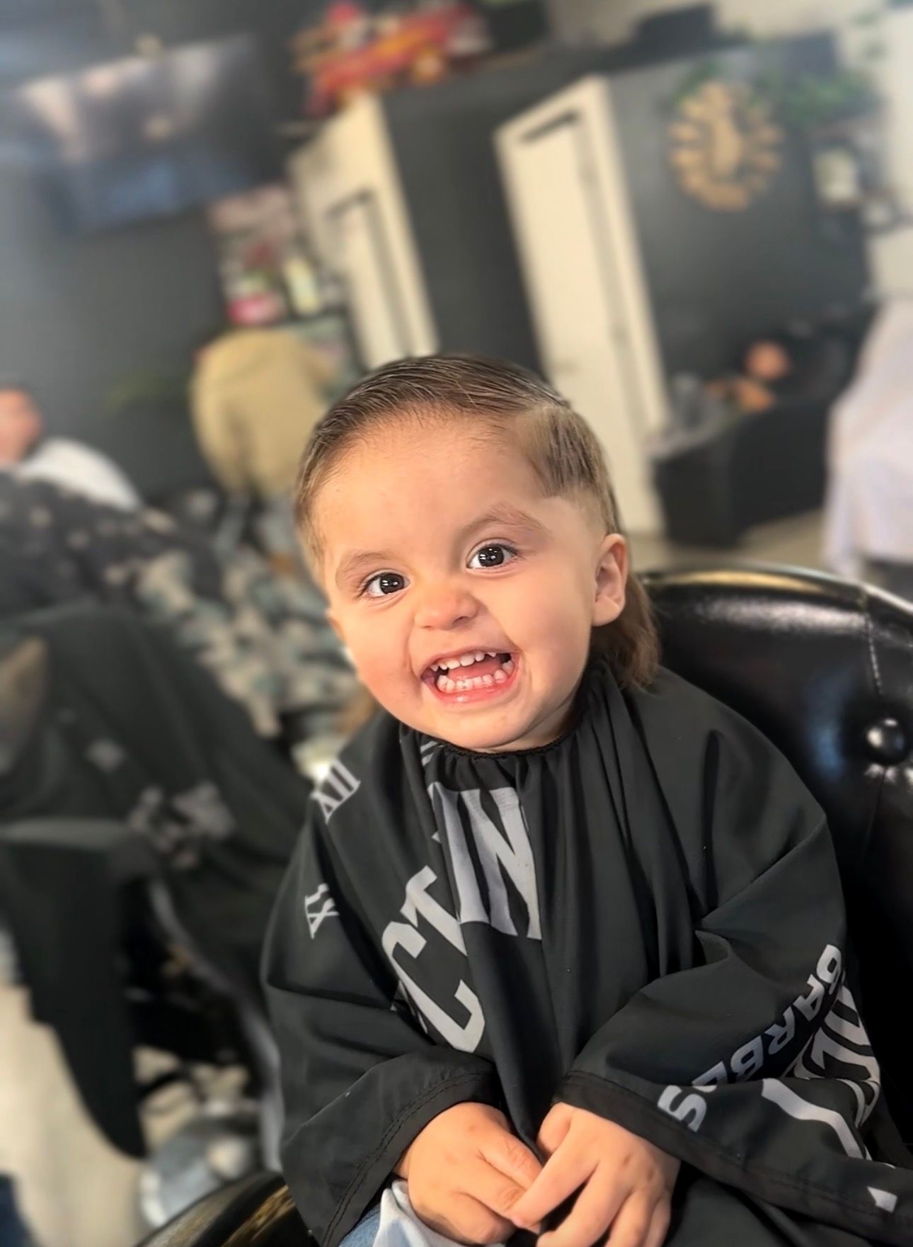 A smiling child in a barber's chair wearing a black cape. They have a new haircut.