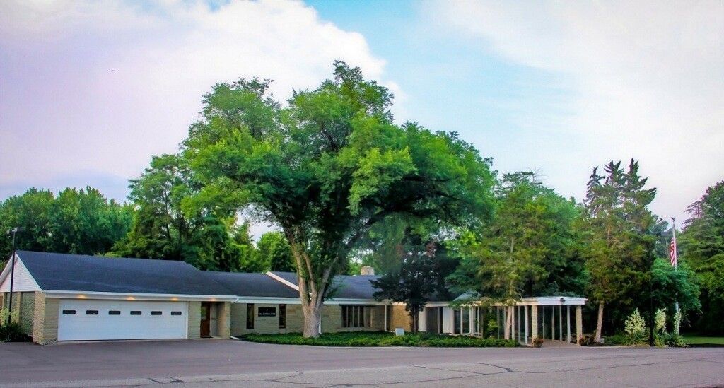 Single-story building with a dark roof and large trees in front. Cloudy blue sky.