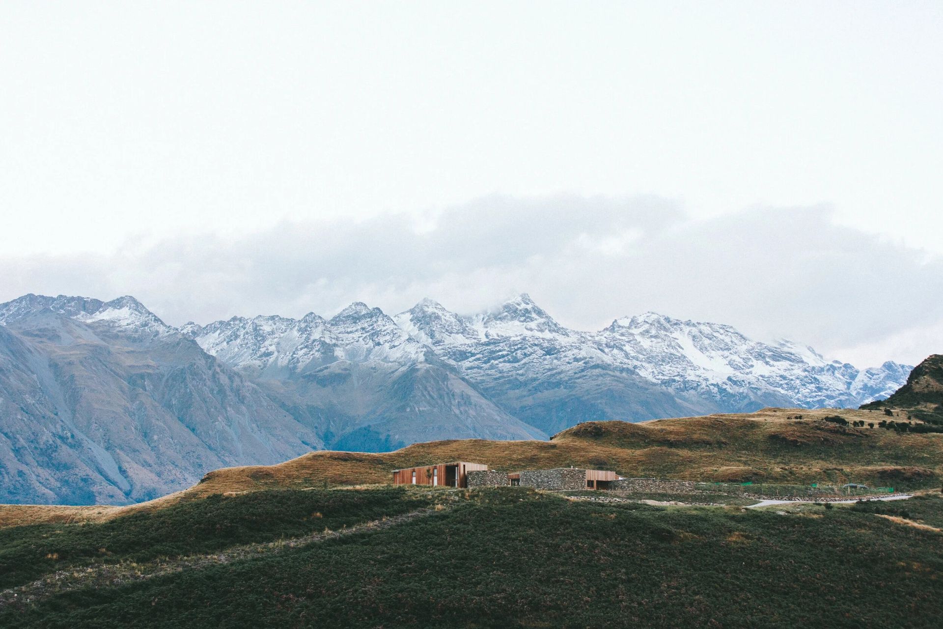 Snowy mountains behind a grassy hillside with a small building under a cloudy sky