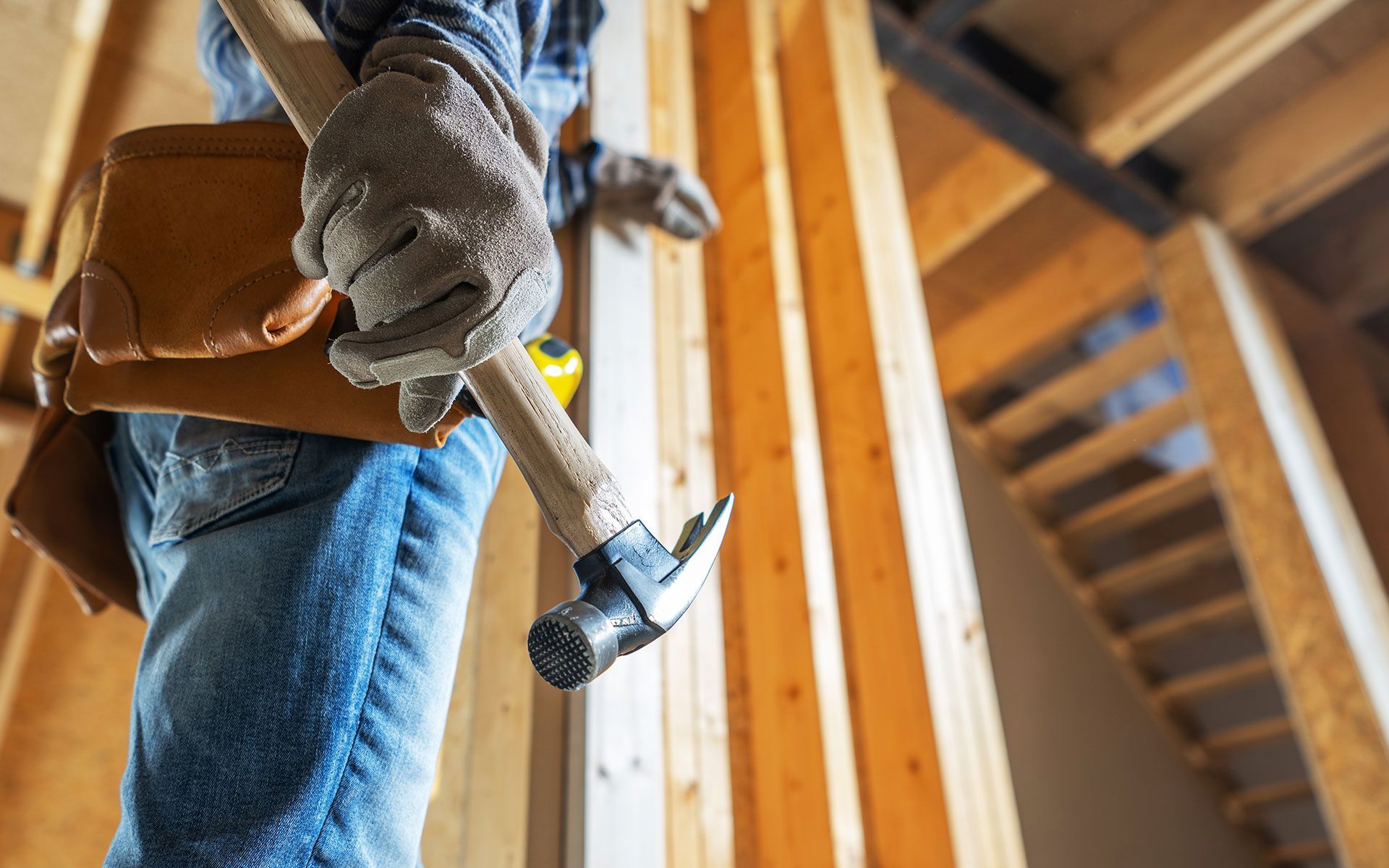 Construction worker holding hammer inside a wooden framed building, wearing gloves and a tool belt.