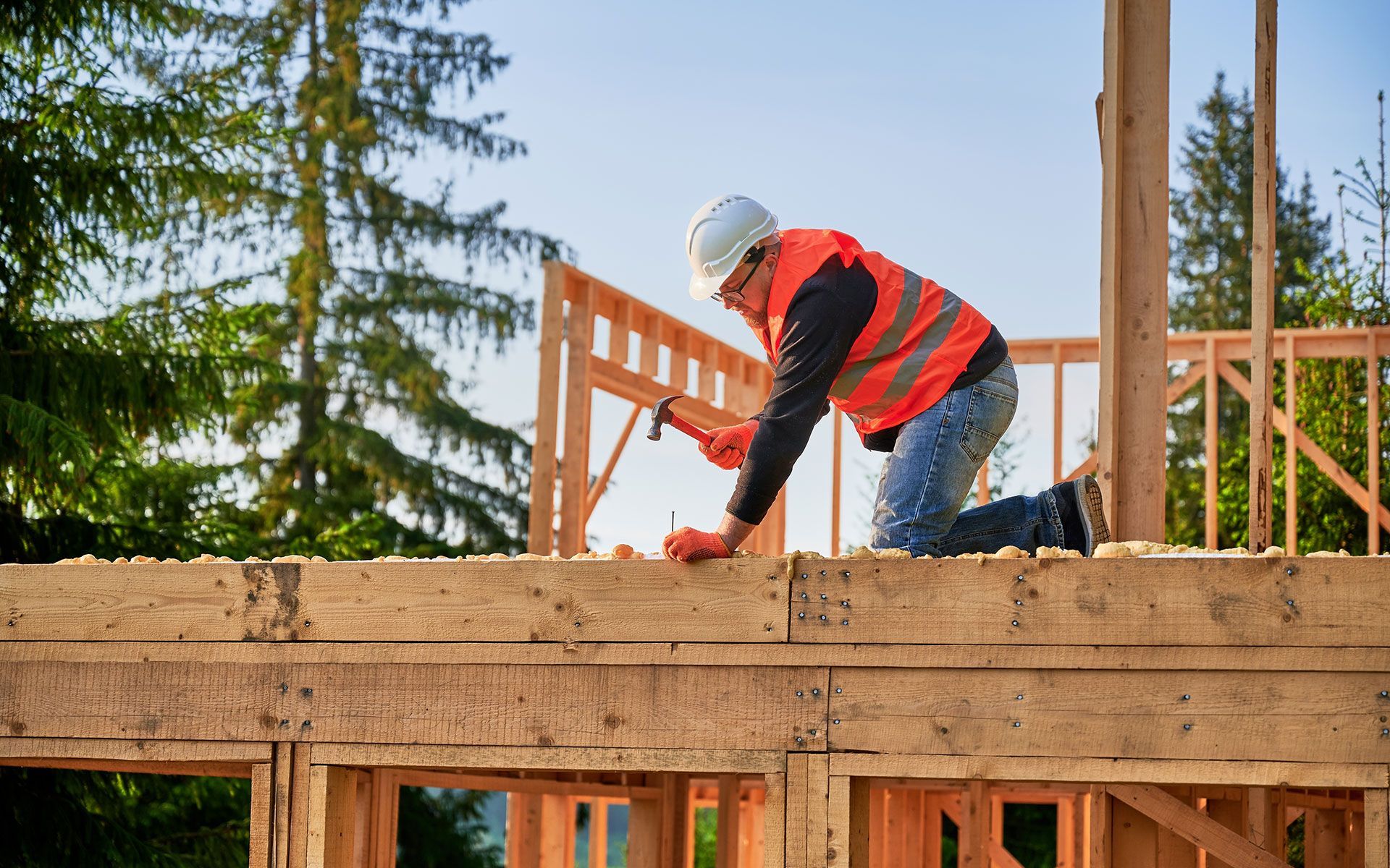 Construction worker hammering on a wooden frame at a building site, wearing safety gear.