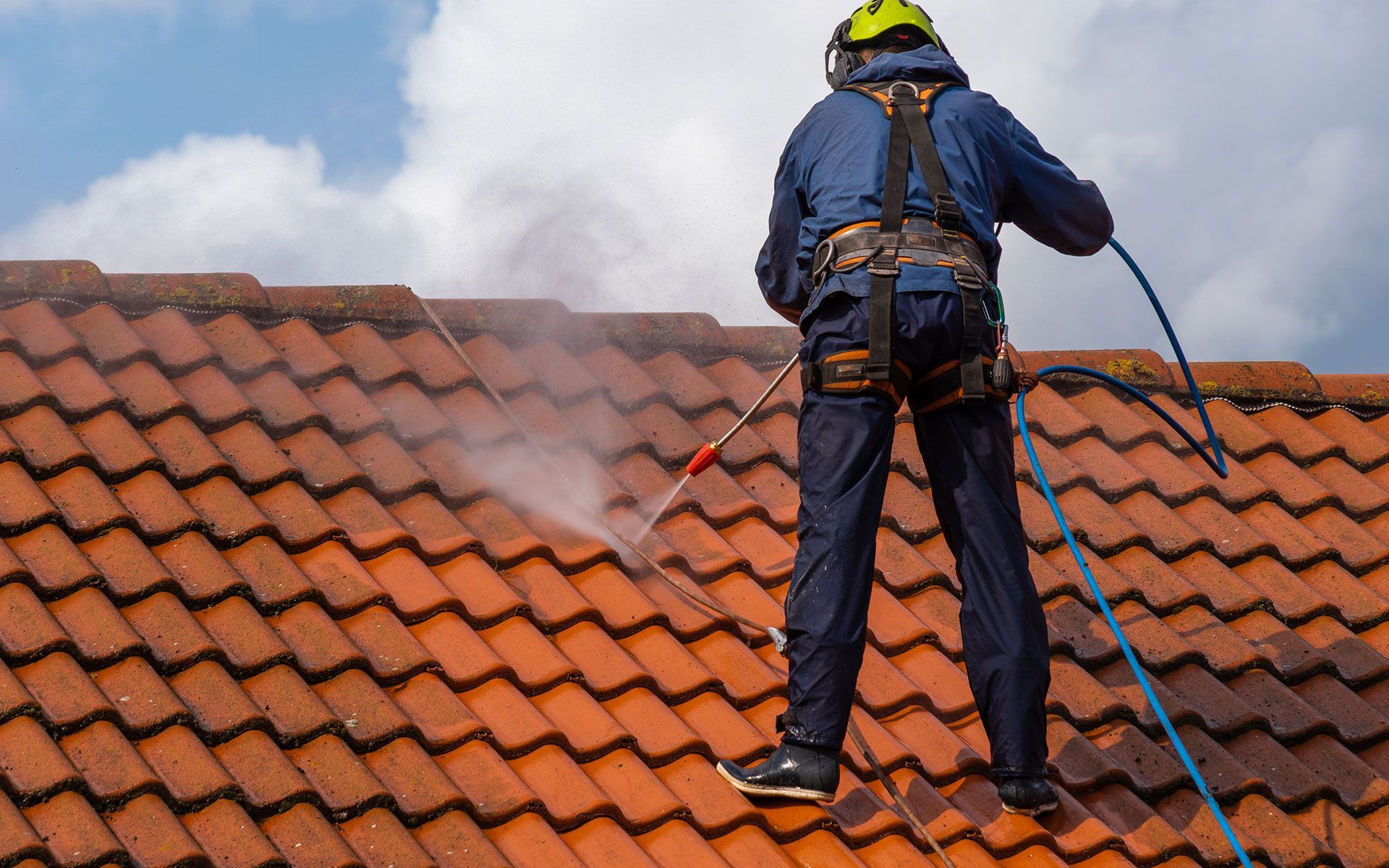 Person in harness power washing a red tile roof on a cloudy day.