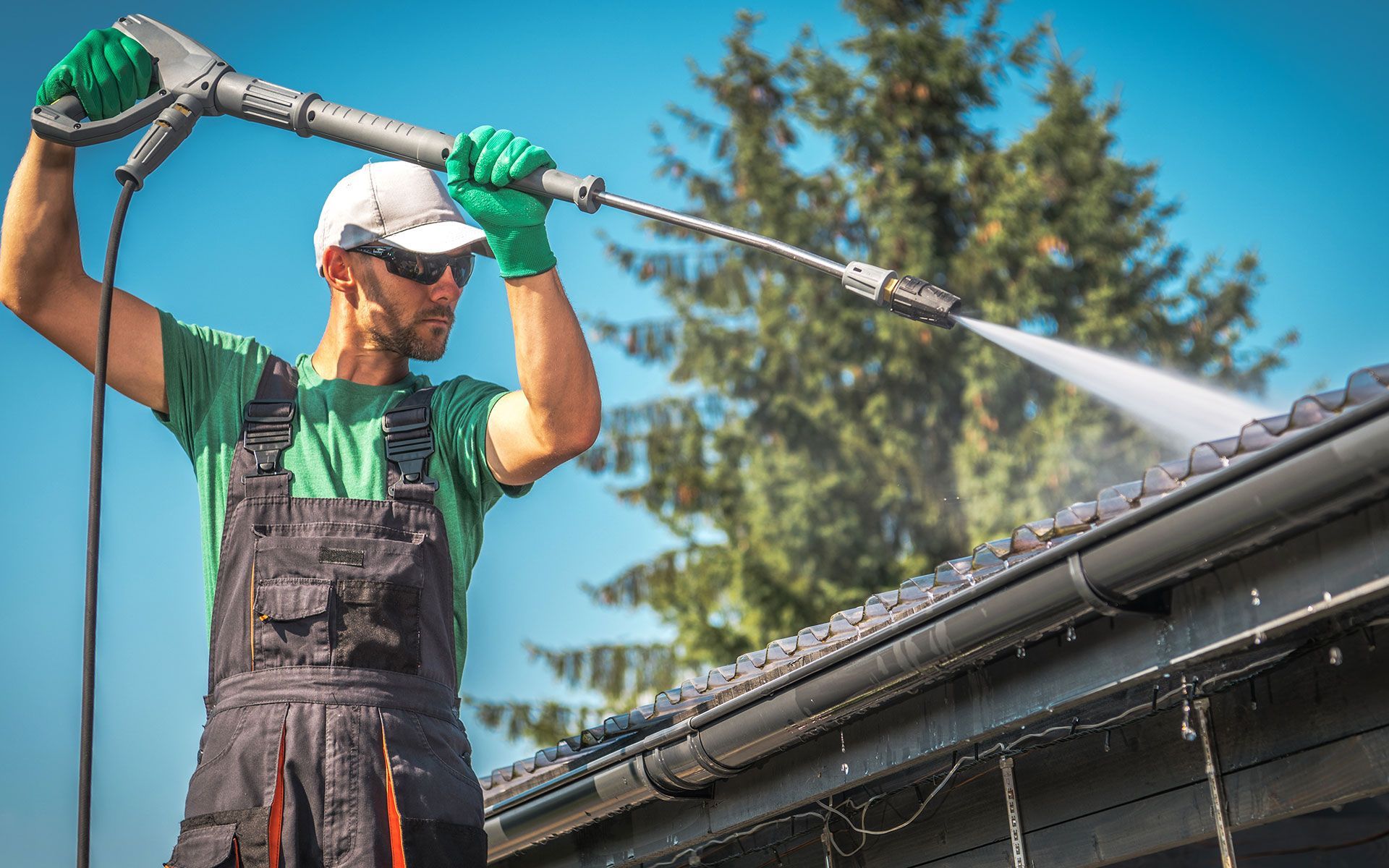 Man pressure washing a gutter on a roof, wearing overalls, gloves, and a hat, on a sunny day.