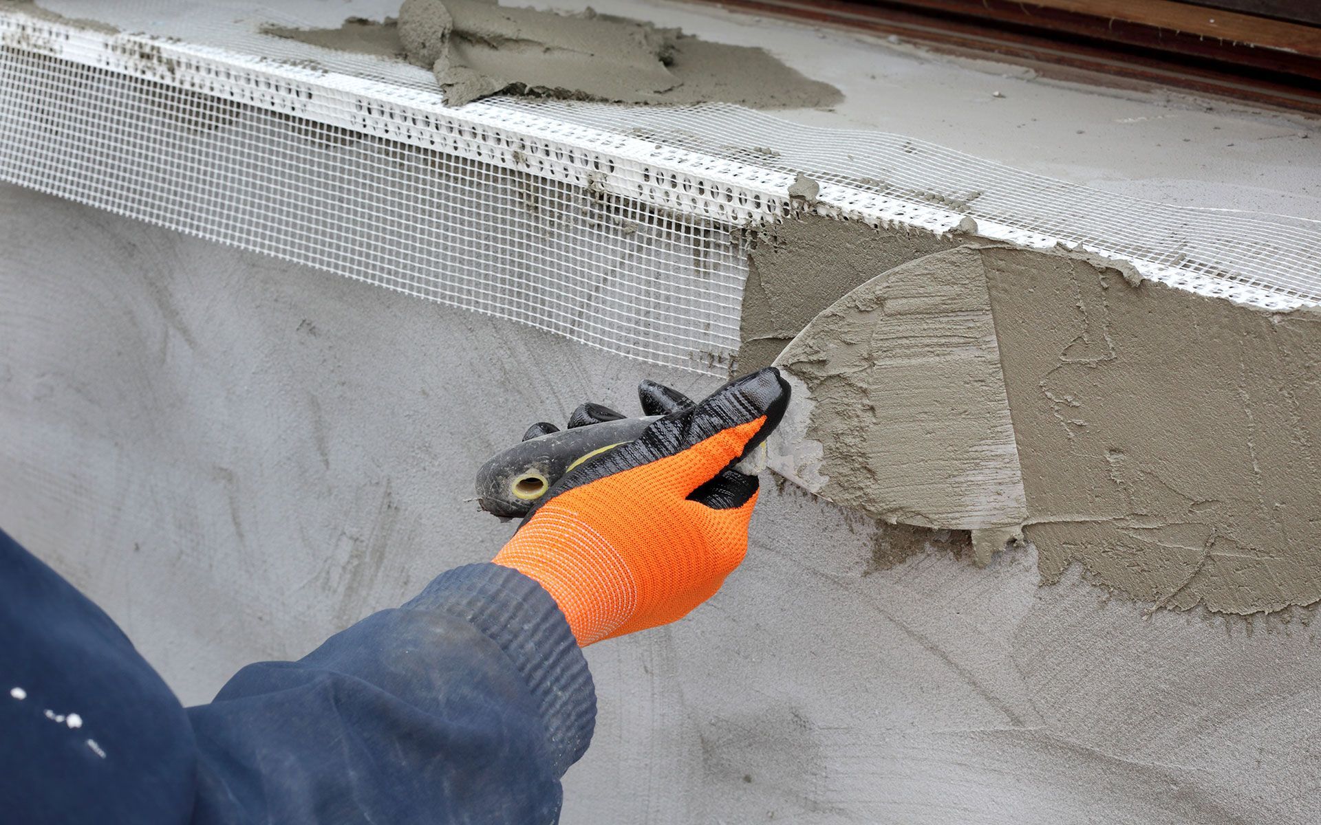 Person with orange glove applies cement to exterior wall with a trowel.