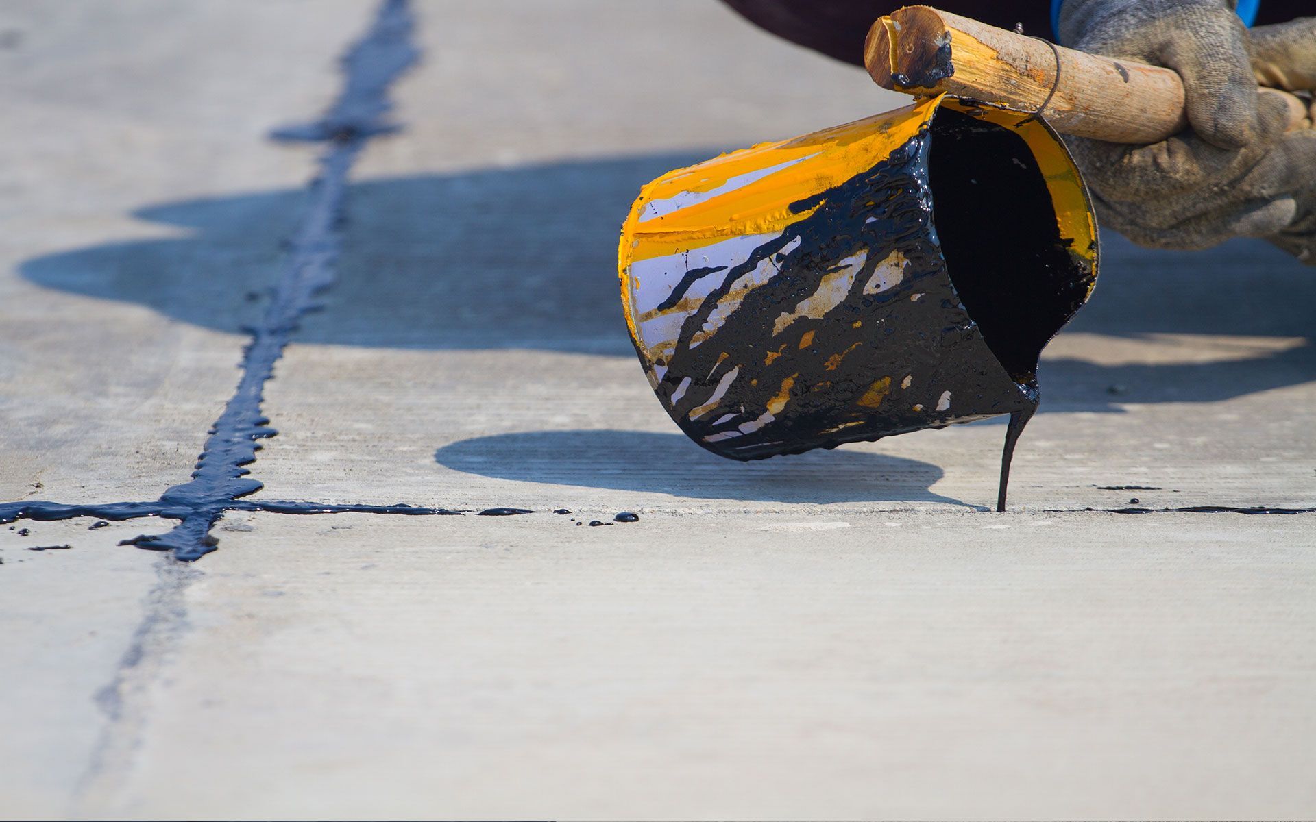 Person pouring black sealant from a yellow bucket into a concrete joint.
