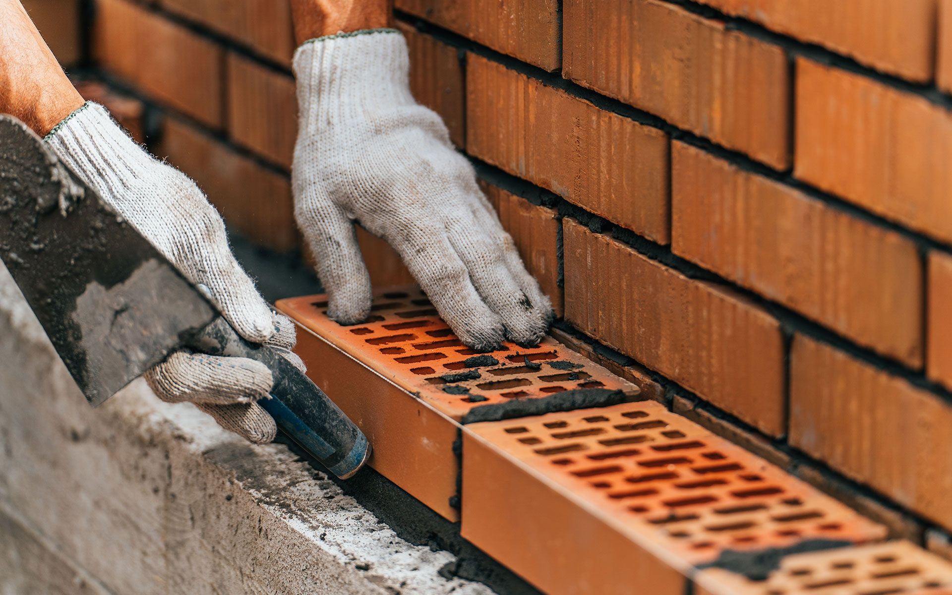 A worker wearing gloves uses a trowel to lay brick, building a wall. Close-up on hands and bricks.