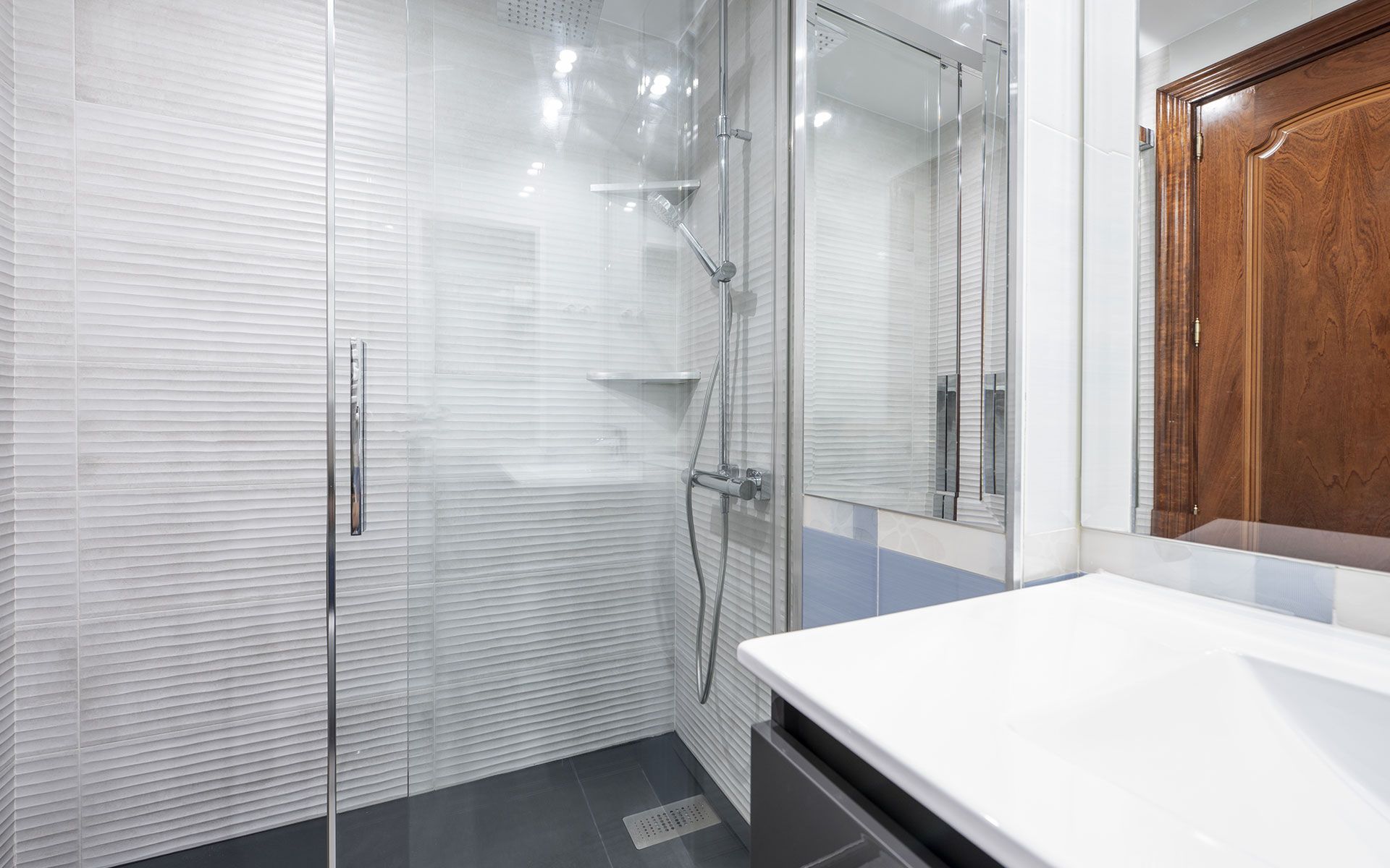 Modern bathroom with glass shower, white countertop, dark floor, and wooden door.