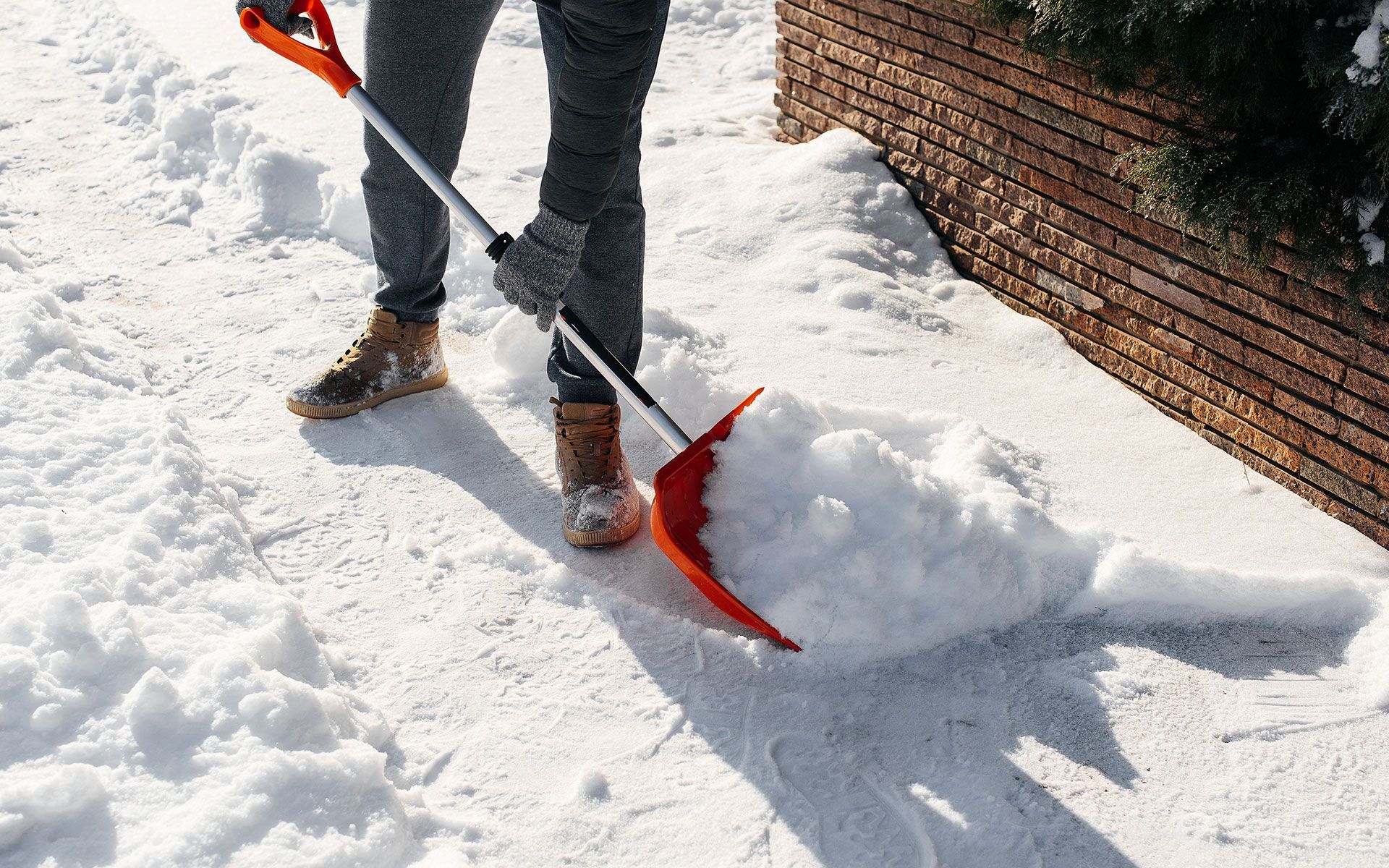 Person shoveling snow off a sidewalk with an orange shovel; a brick wall is visible.