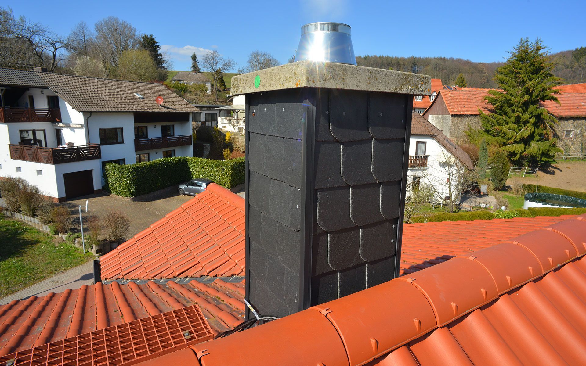 Chimney on a red tiled roof with slate siding and metal cap; houses in background.
