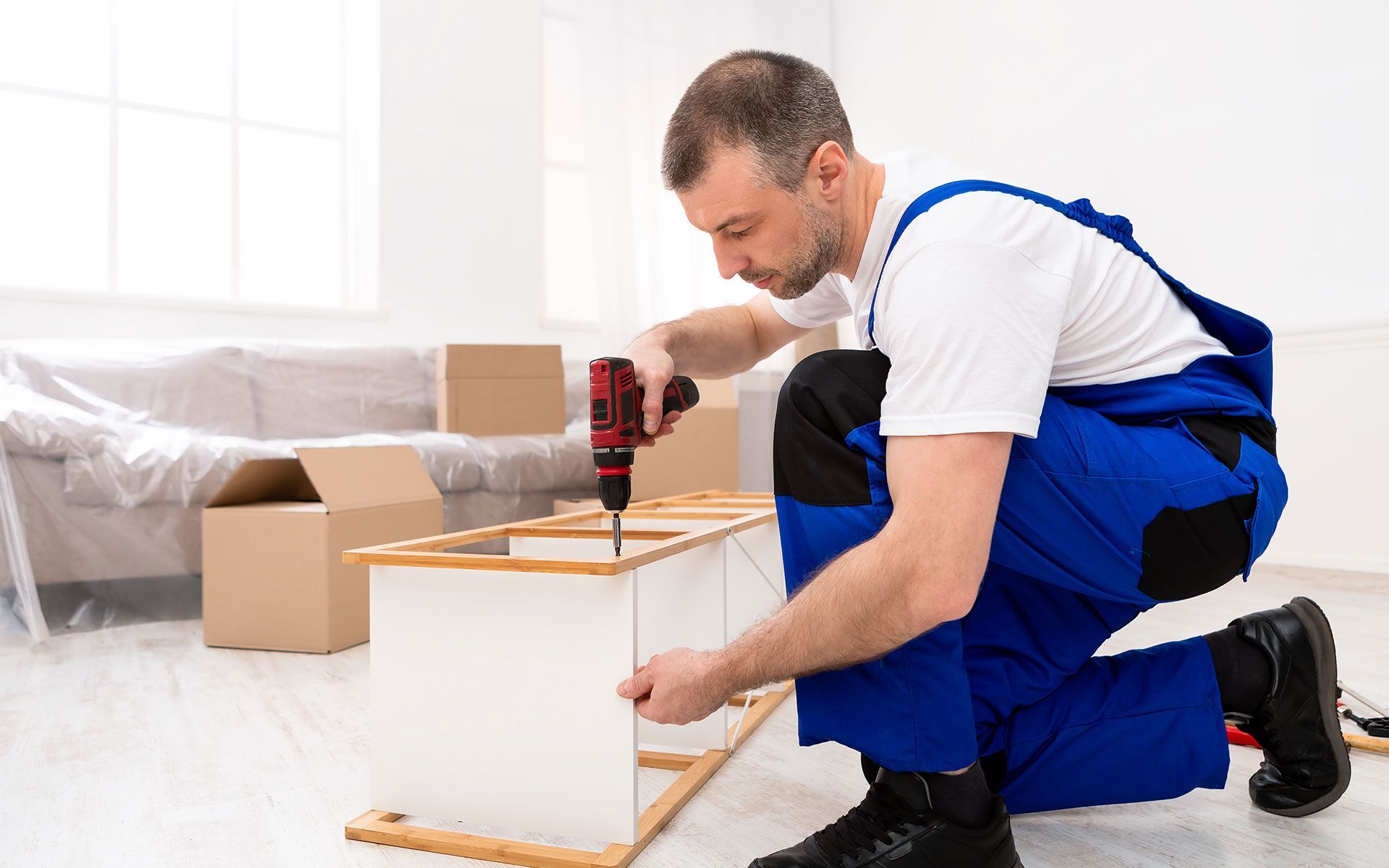 Man in blue overalls assembling furniture, using a power drill indoors.