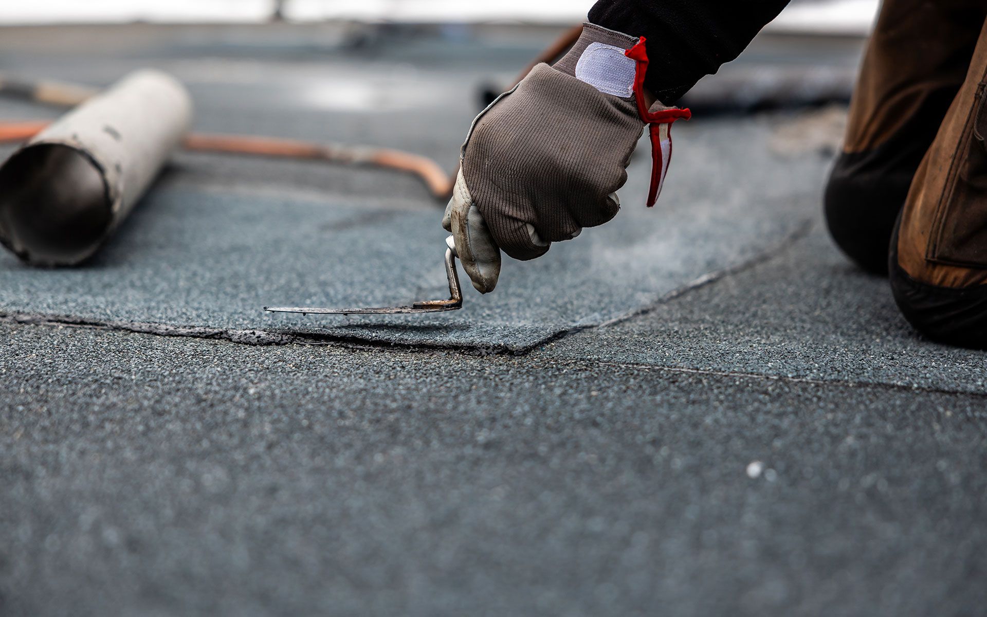 A worker repairing a dark roofing material with a tool, wearing gloves, close-up.