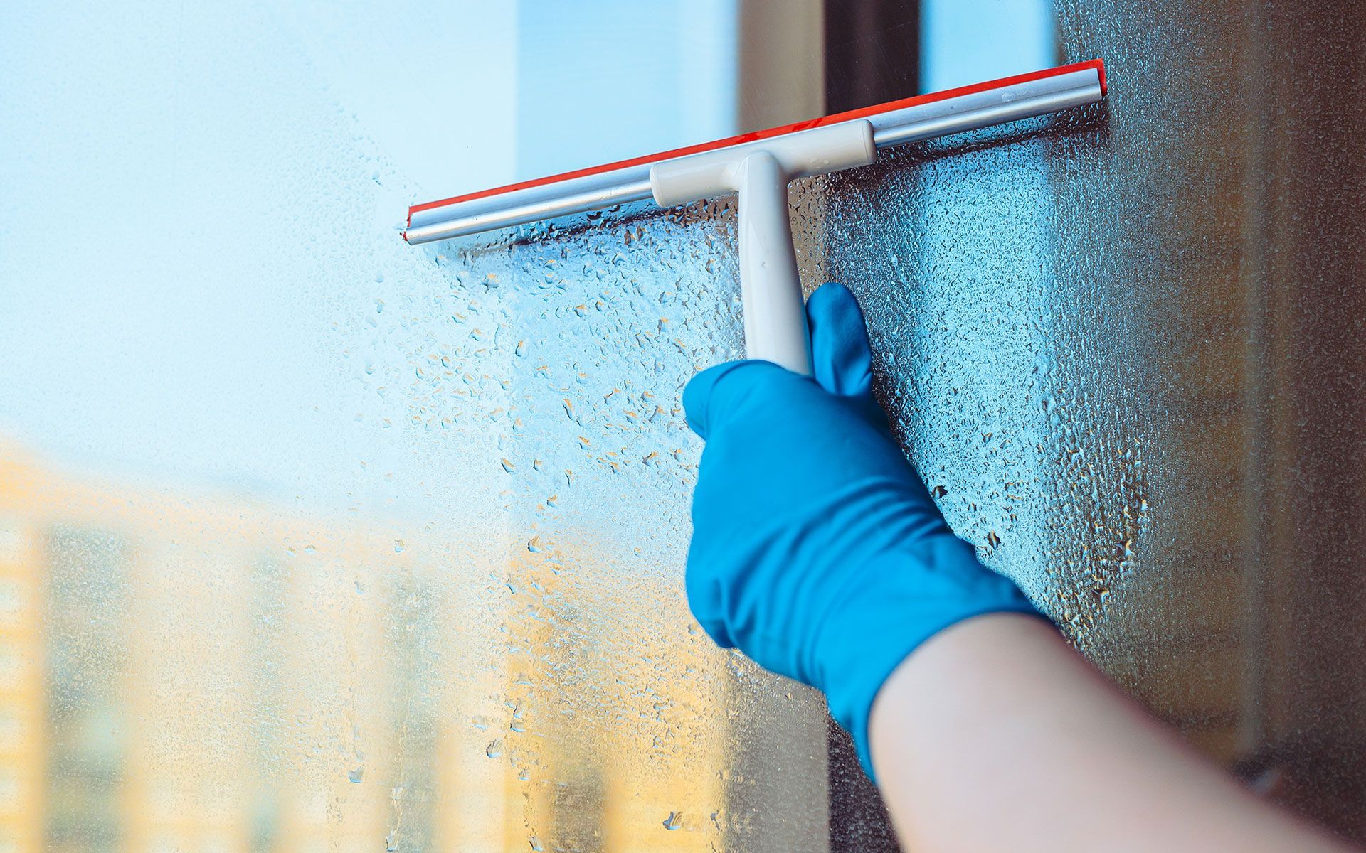Hand in blue glove using squeegee to clean a window with condensation.