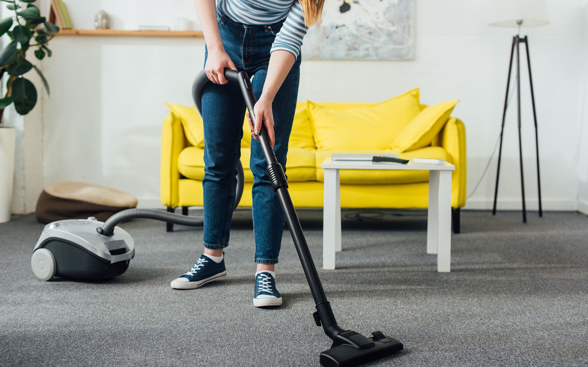 Woman in jeans vacuums a gray carpet in a living room with a yellow couch.