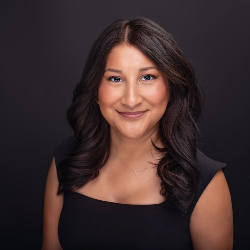 Woman with dark hair and a black dress smiles at the camera against a black background.