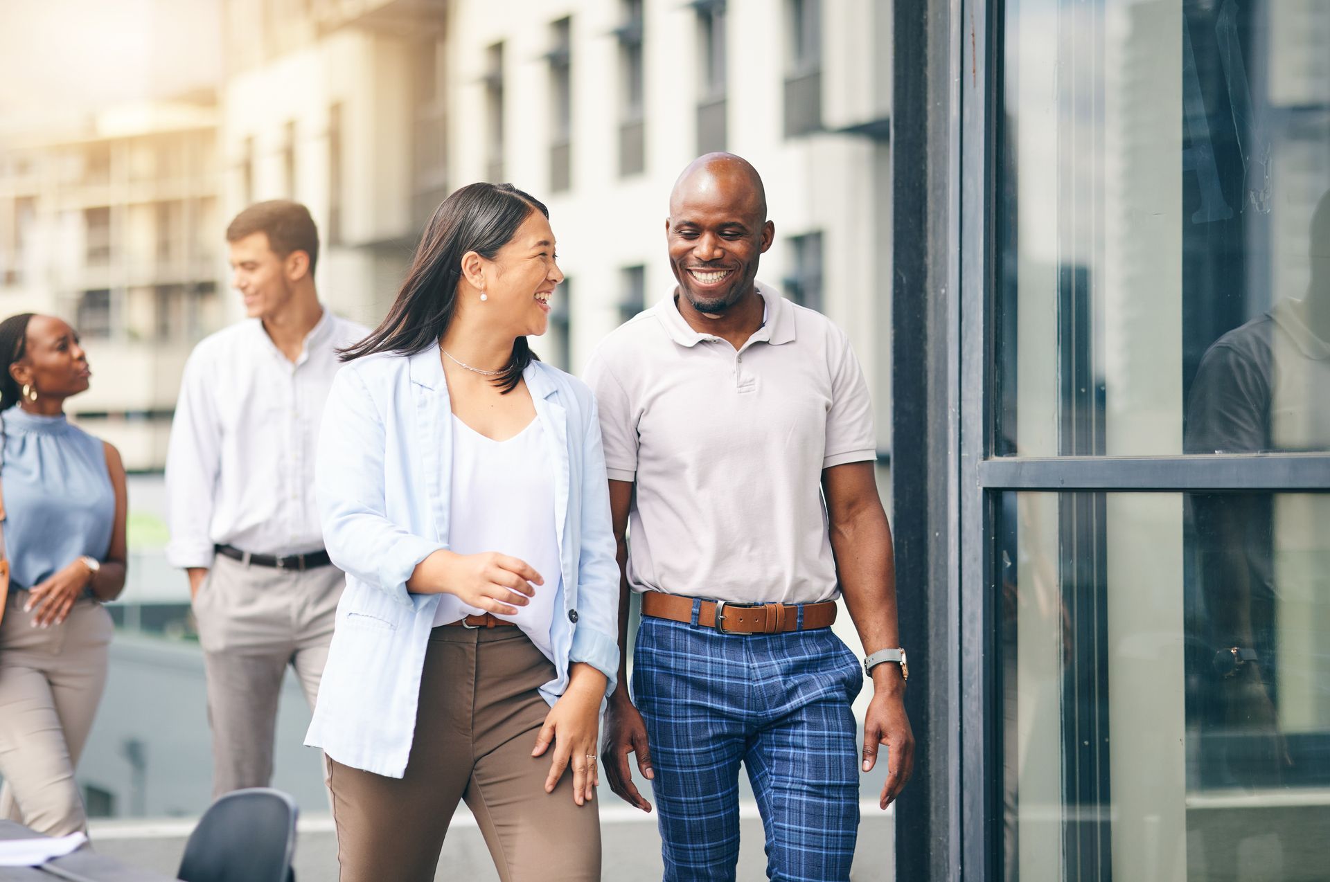 Two smiling colleagues walking and talking on an office balcony; other professionals visible.