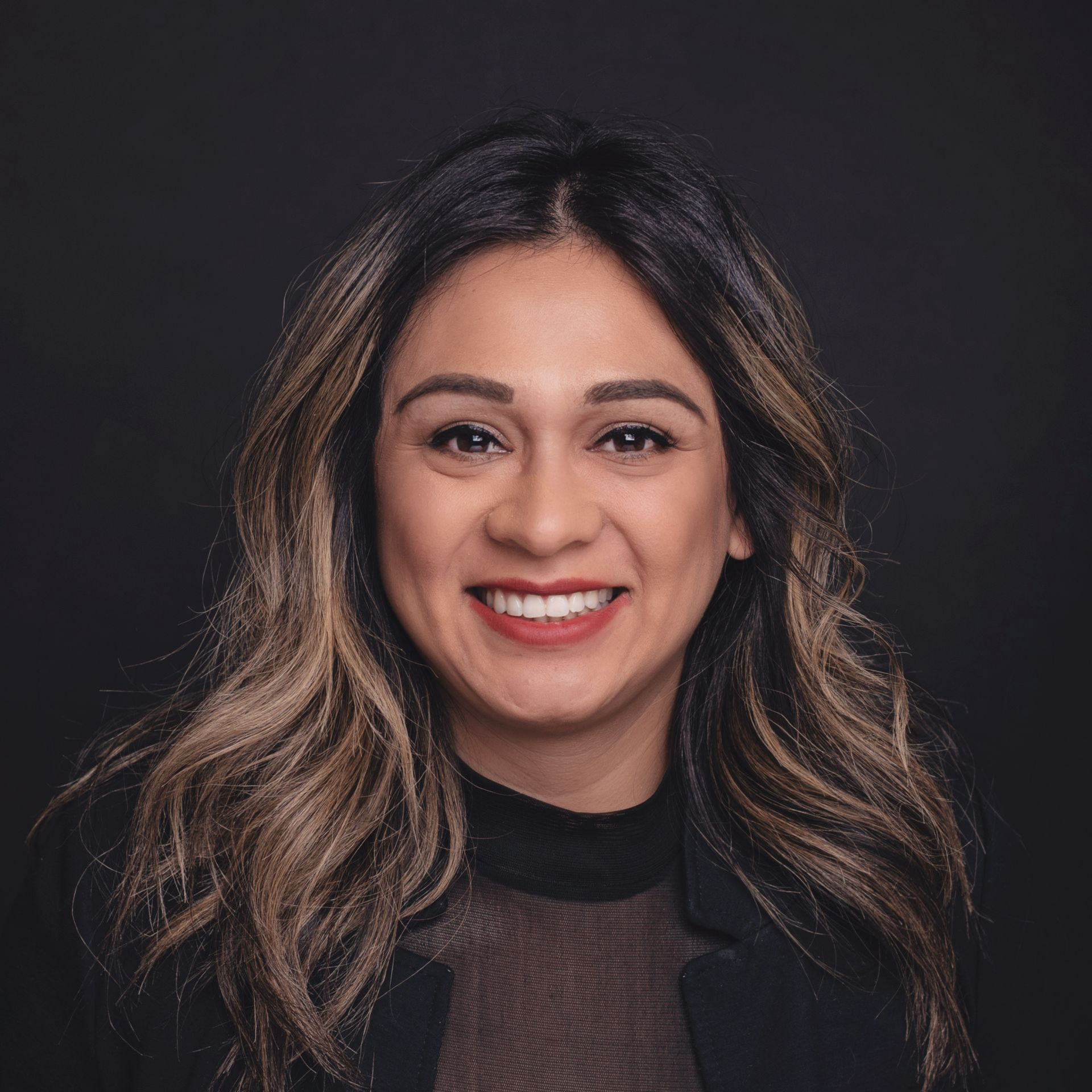 Woman with dark hair and highlighted tips smiles at the camera against a dark backdrop.
