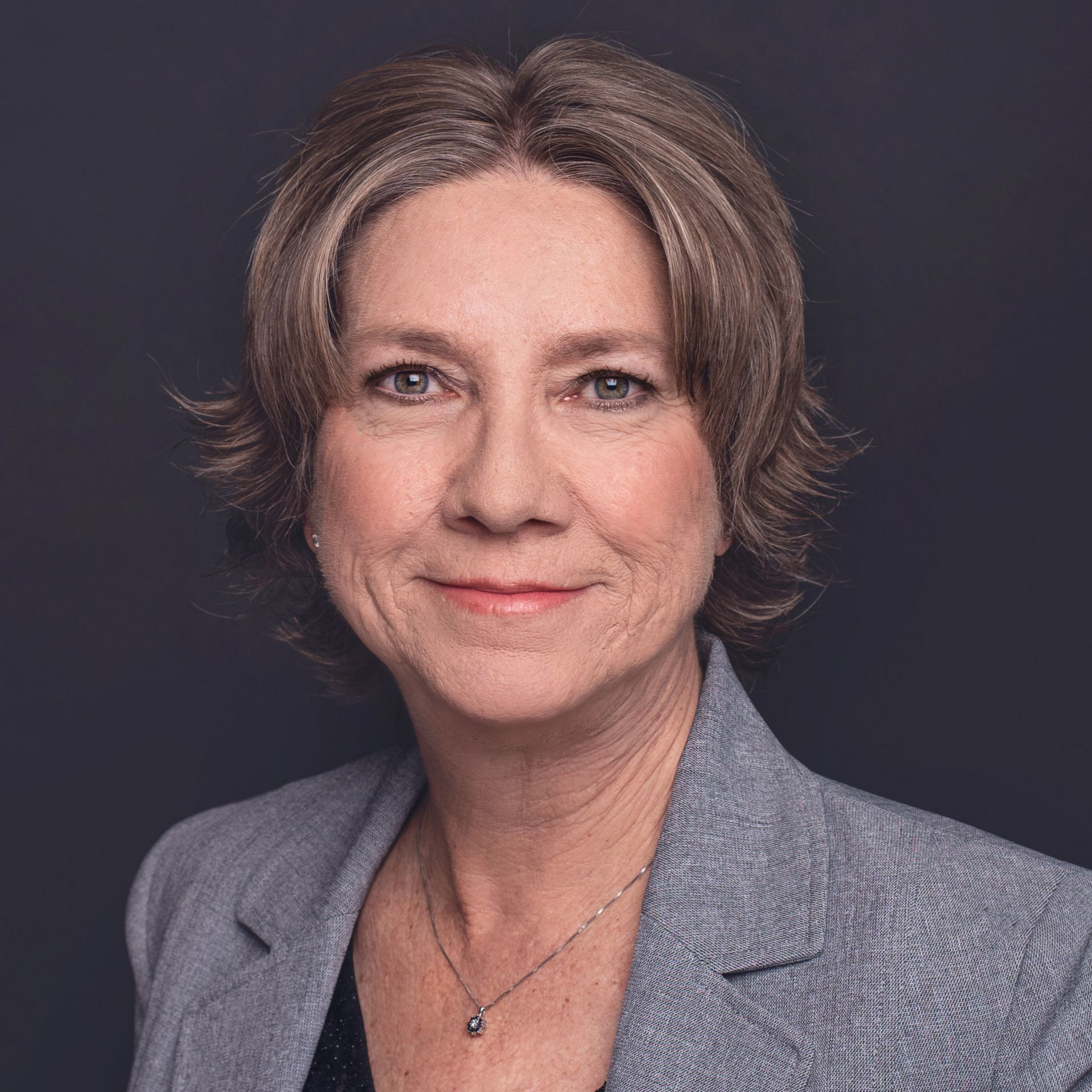 Woman with short, graying hair, smiling, wearing a blazer and necklace against a dark background.
