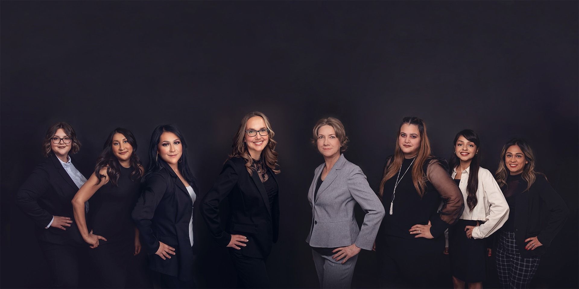 Group of professional women in suits, hands on hips, against a dark background.