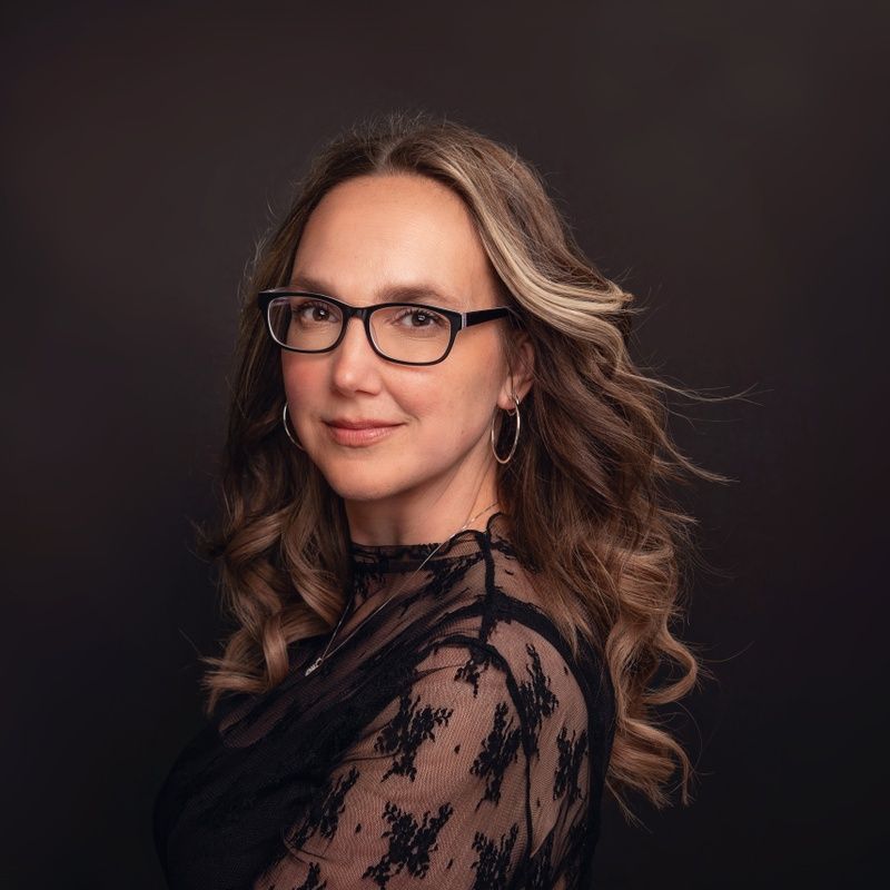 Woman with glasses, wearing a black lace top, smiles at the camera. Against a dark backdrop, long wavy hair.