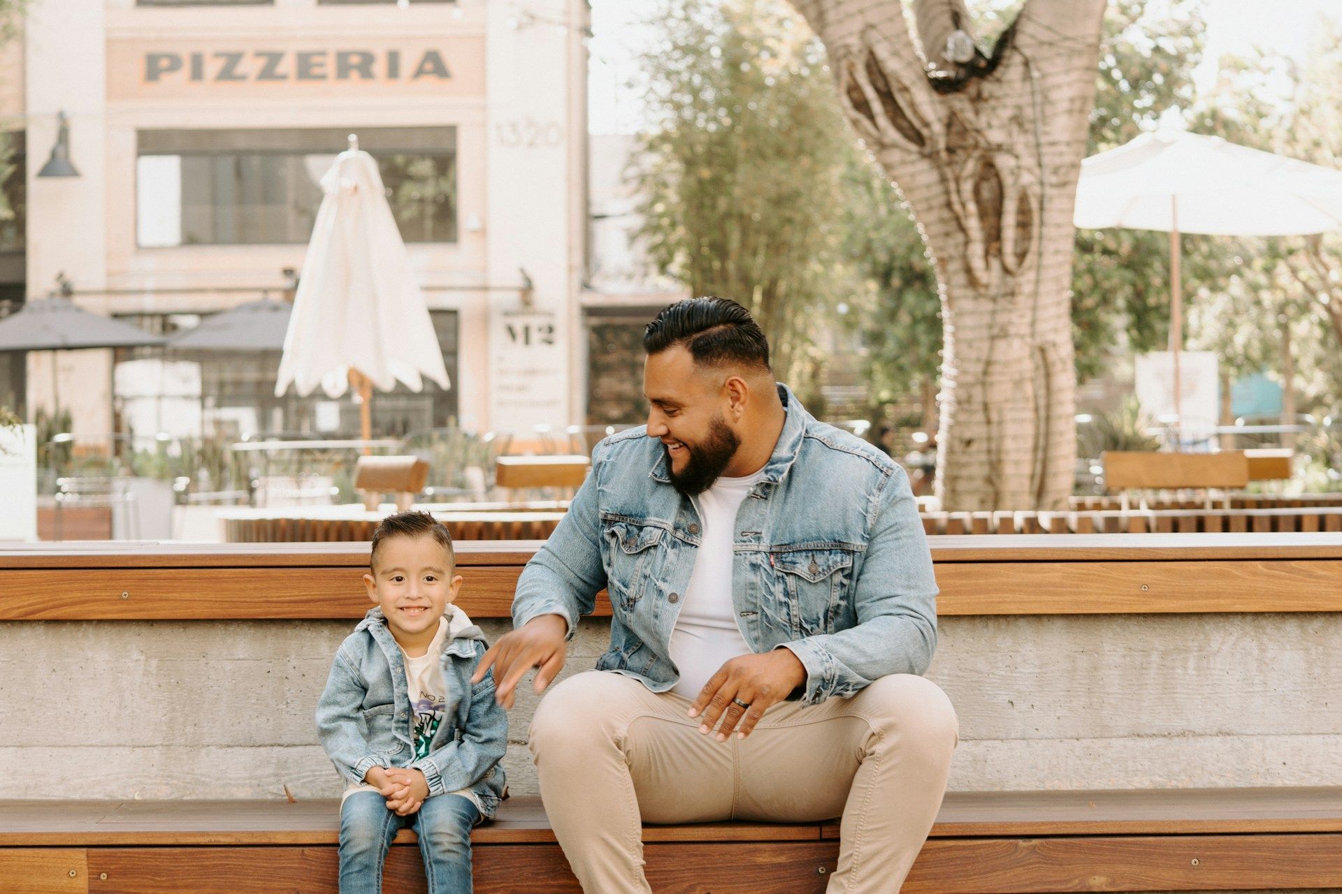 Father and child in matching denim jackets sitting on a wooden bench outside a pizzeria.