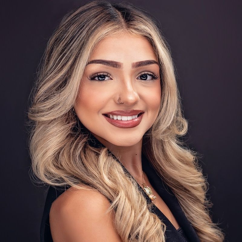 Smiling woman with wavy blonde hair, dark top, and nose ring, against a dark background.