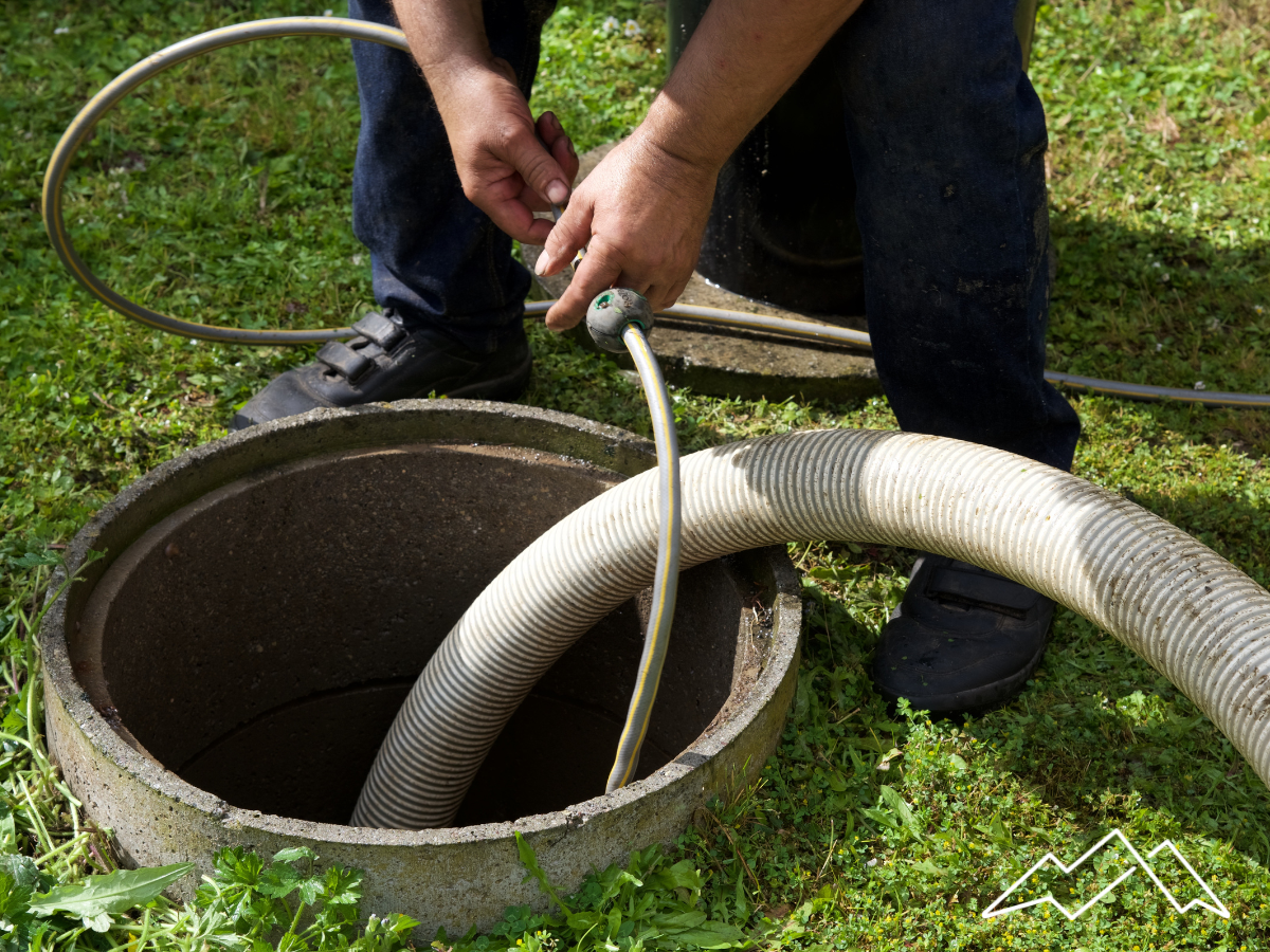 A person in jeans and shoes with a hose inserted into a concrete septic tank in a grassy yard.