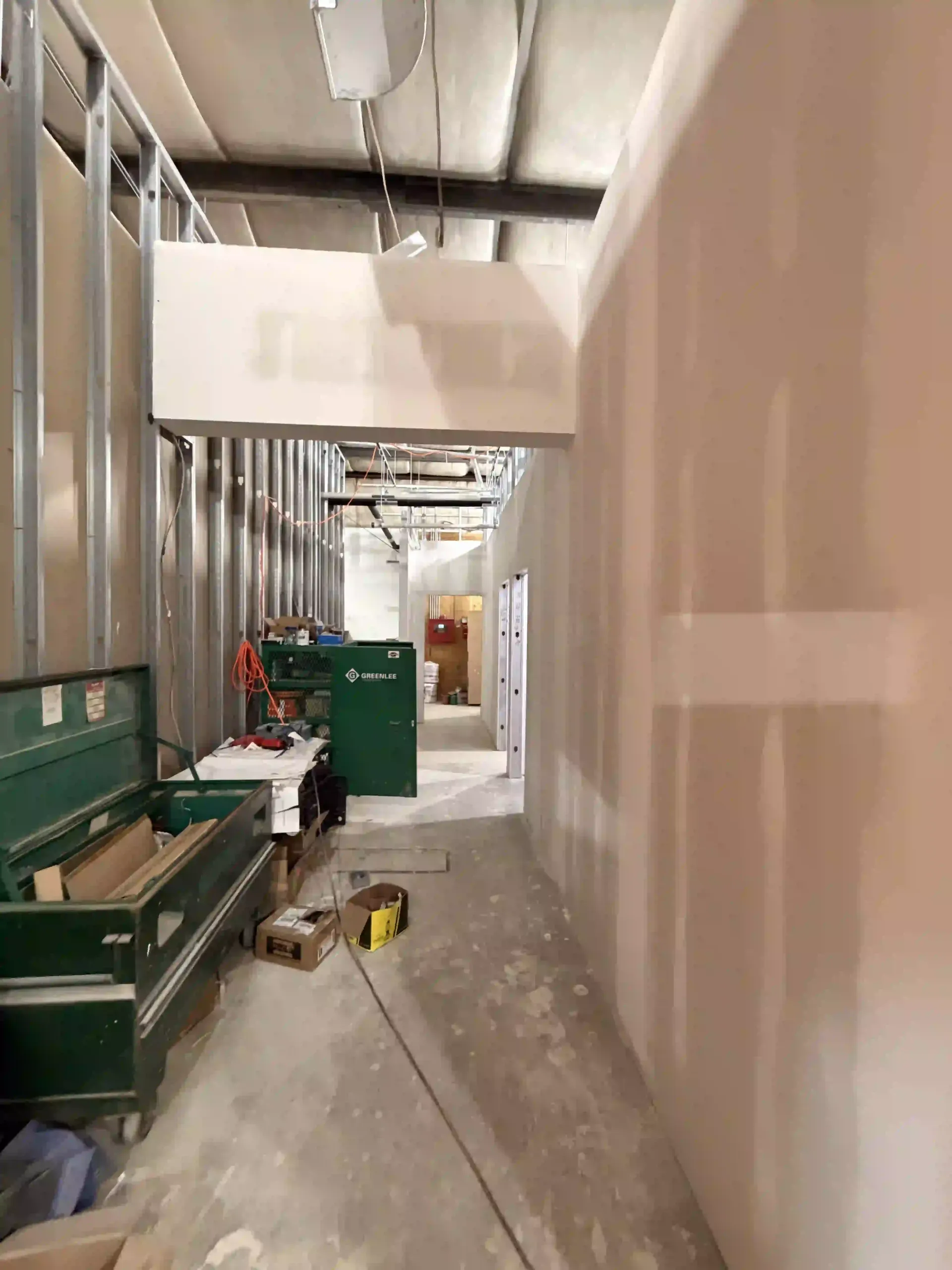 Narrow construction corridor with unfinished walls, green equipment, and tools along a dusty floor