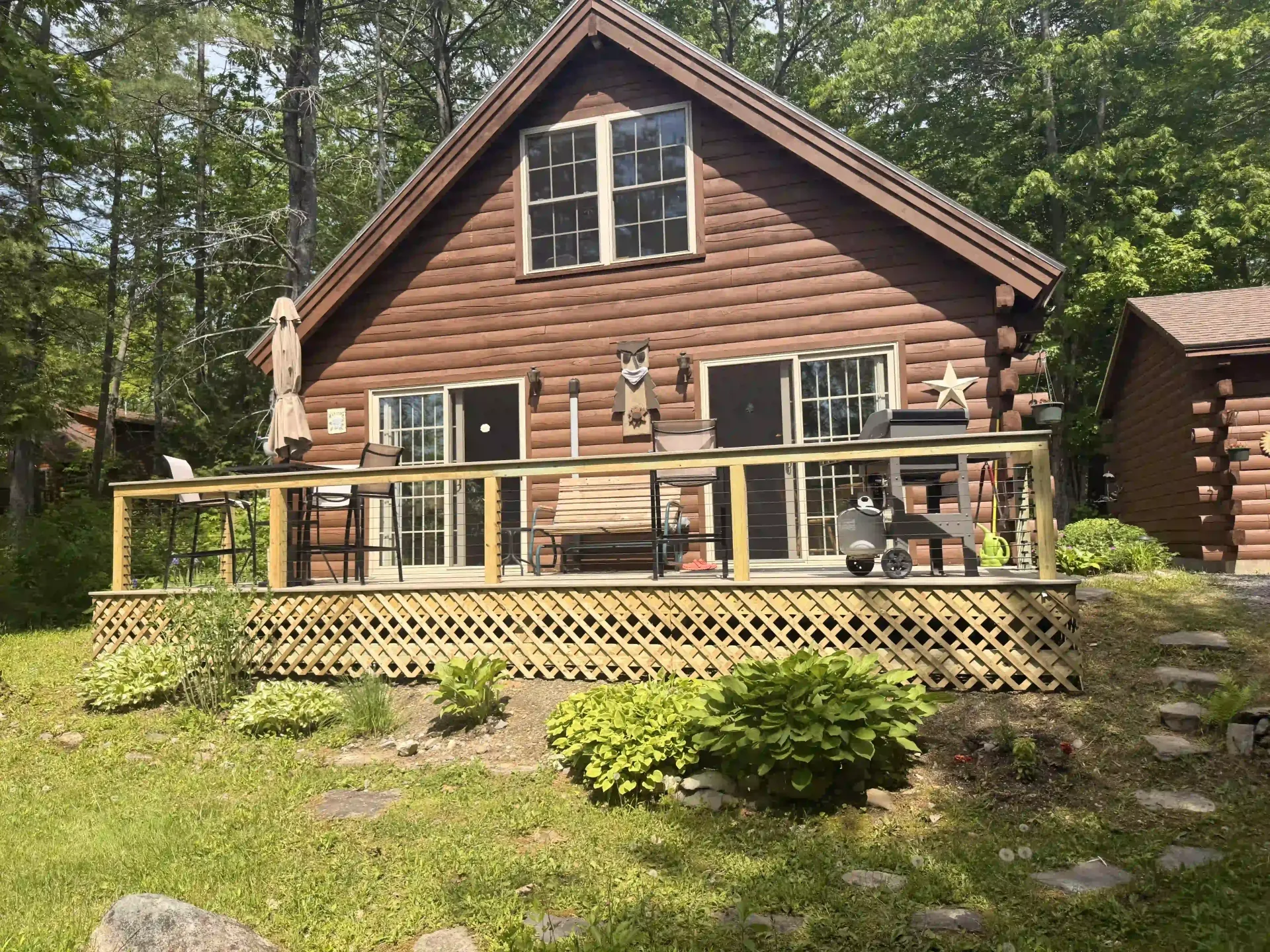 Log cabin with a deck and railing, surrounded by green trees and shrubs.