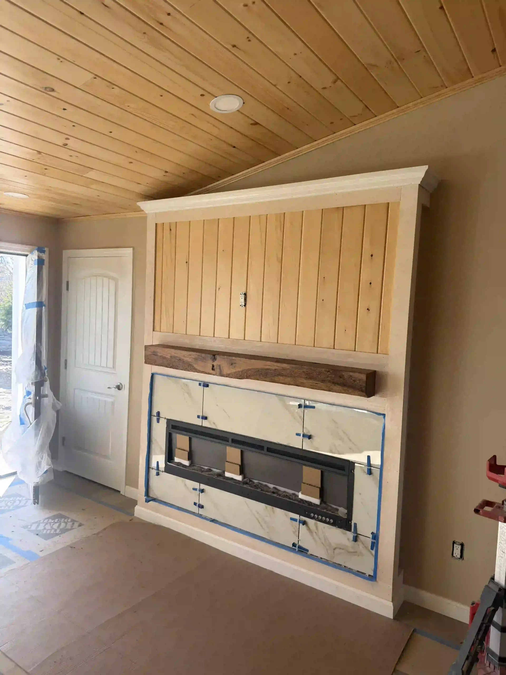 Wood-paneled ceiling room with a built-in wall unit, open cabinet, and a fireplace insert under construction