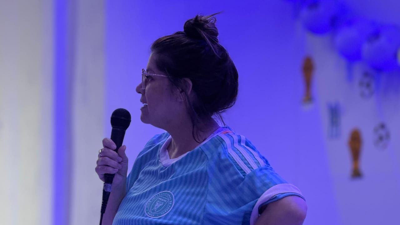 Mujer cantando con micrófono, con el brazo levantado en señal de adoración. Un guitarrista al fondo. Interior con iluminación tenue.