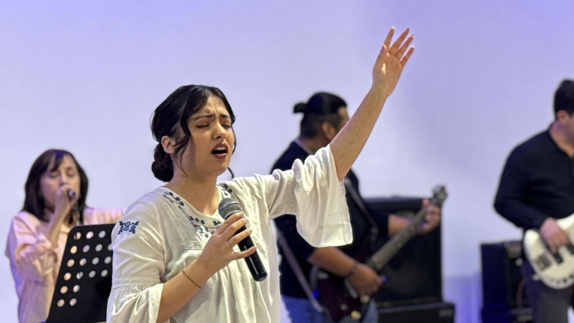 Mujer cantando con la mano levantada, con banda tocando en el escenario. Camisa blanca, fondo azul.