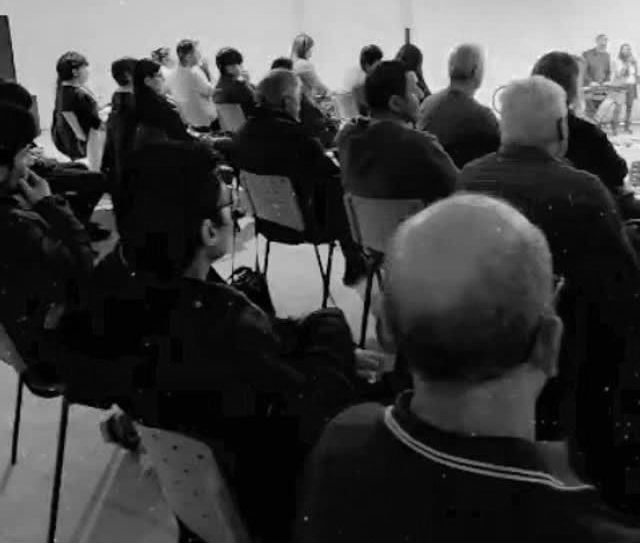 Público sentado frente a un escenario con un fondo para presentaciones. Iluminación tenue; la gente escucha.