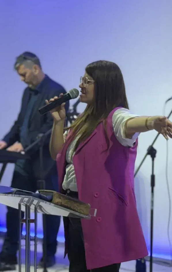 Mujer cantando frente a un micrófono, vistiendo un chaleco rosa, con un hombre tocando un teclado de fondo.
