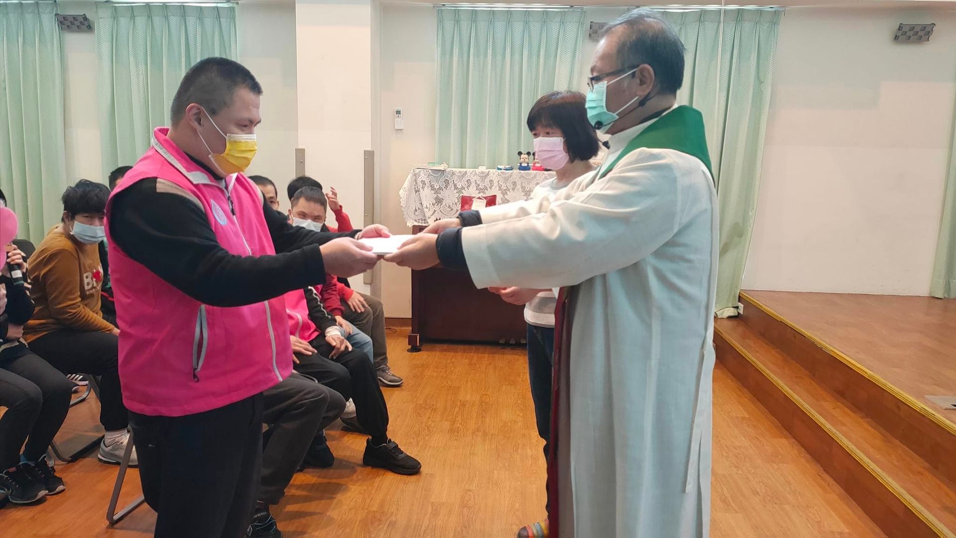 Two people exchanging an item in a hall, one in a pink vest and one in a white robe.