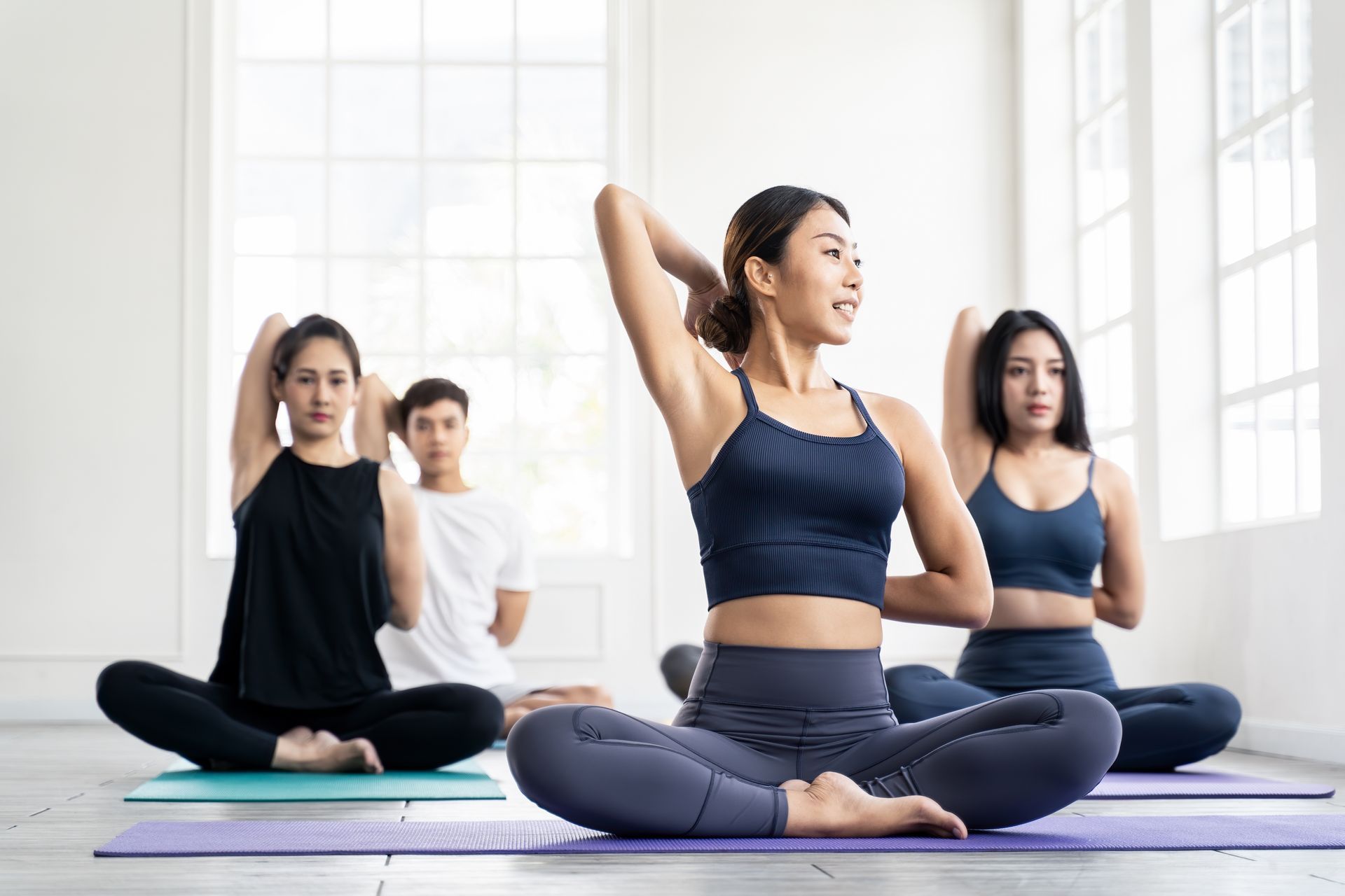 People in yoga class, practicing a seated pose with arms behind the back, indoors in a bright room.