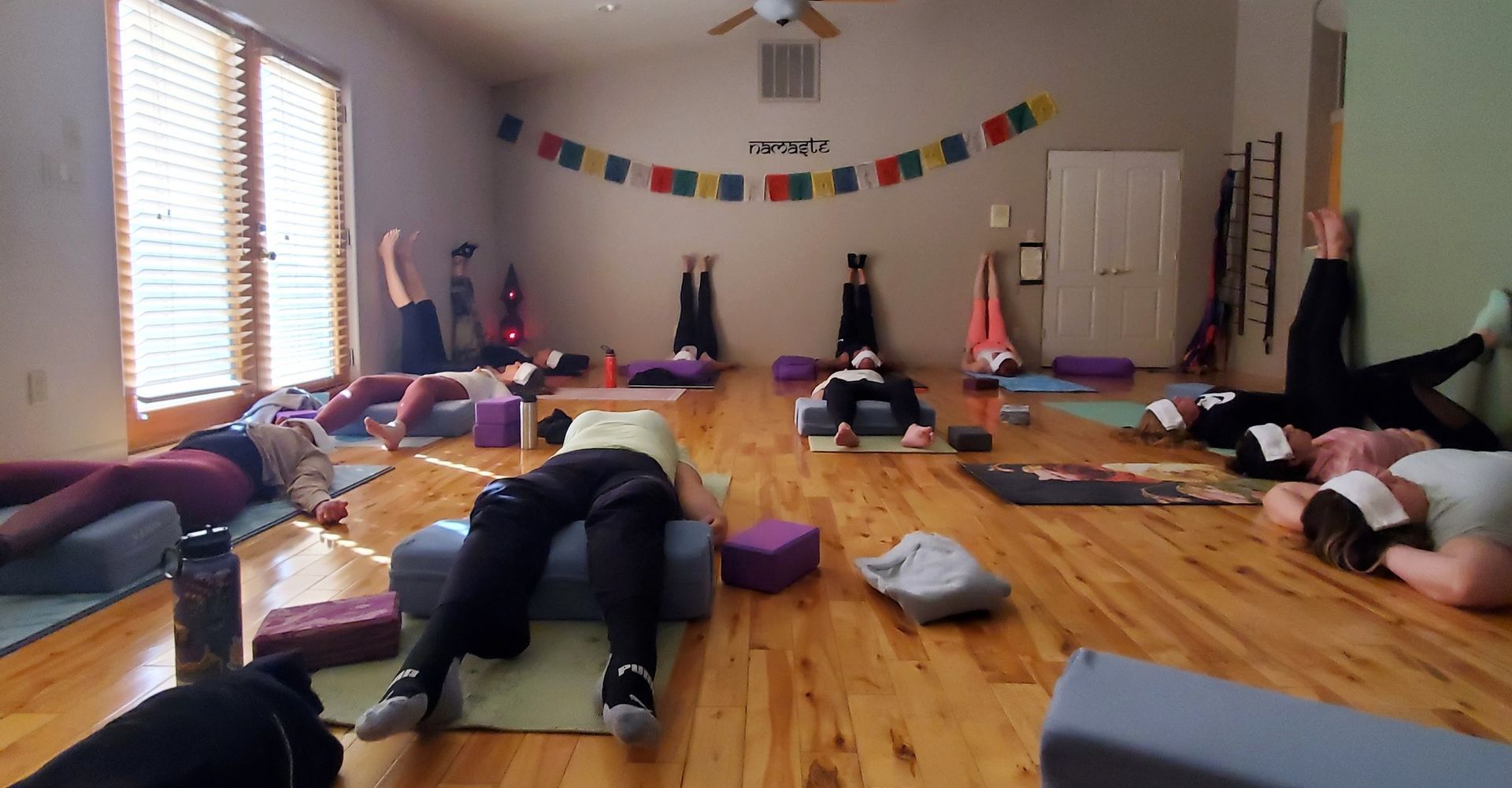 People in a yoga class, resting on mats with props, near windows.
