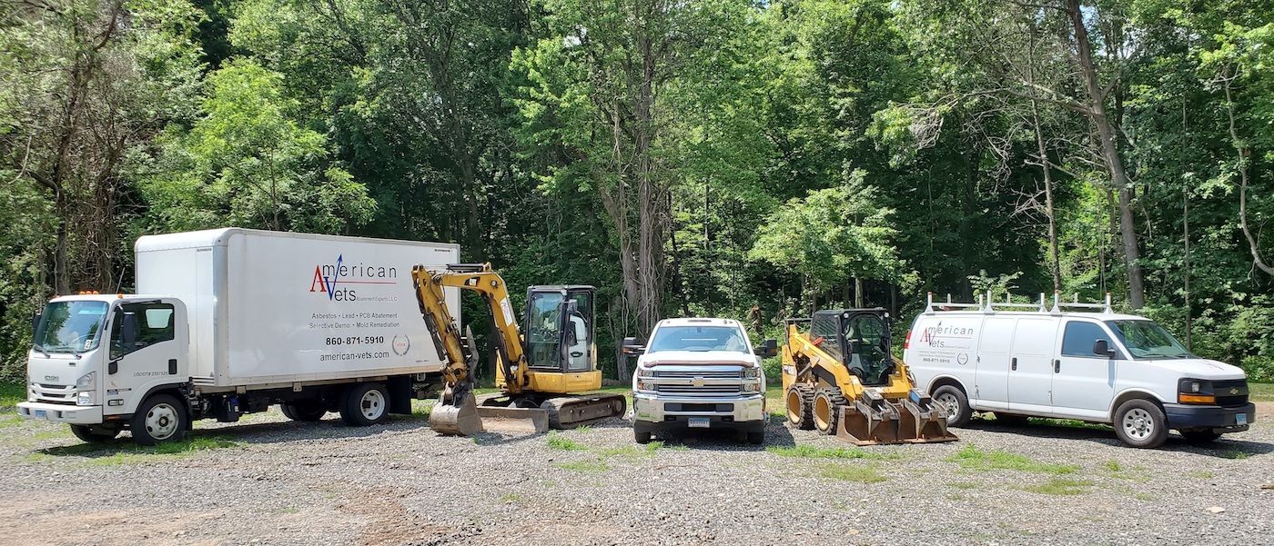A fleet of construction vehicles including trucks, an excavator, and a loader, parked on gravel in front of trees.