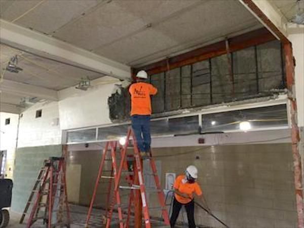 Construction workers removing a wall on ladders, wearing orange shirts and hard hats.