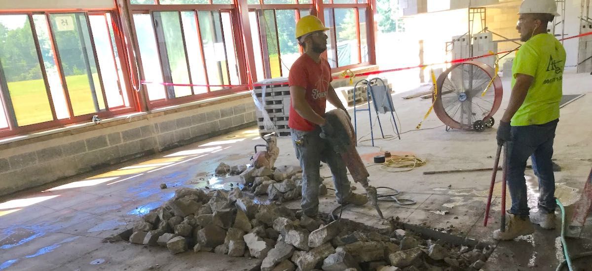 Two construction workers breaking concrete with jackhammer. One wears red shirt and the other wears green. Indoors.