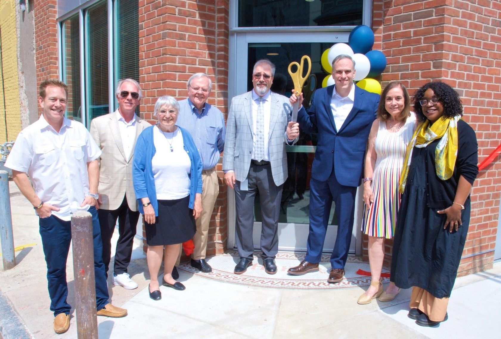 Group of people at a building opening, cutting a ribbon. A man holds oversized golden scissors, balloons, and red ribbon. Group of people at a building opening, cutting a ribbon. A man holds oversized golden scissors, balloons, and red ribbon.