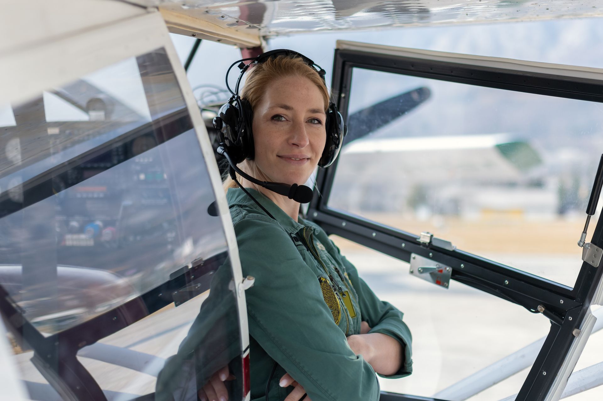 Woman in cockpit wearing a headset, smiling, arms crossed. Airplane interior, airfield background. Woman in cockpit wearing a headset, smiling, arms crossed. Airplane interior, airfield background.