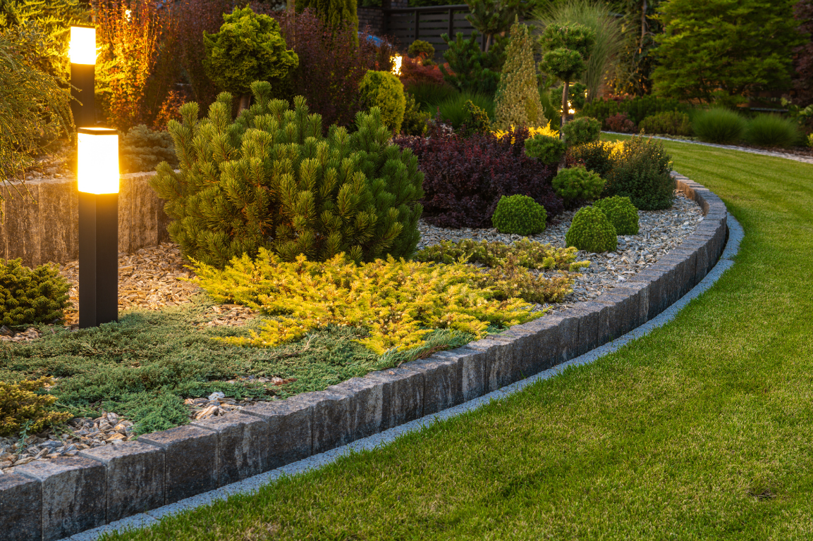 Evening garden with landscape lighting, featuring stone edging, trimmed shrubs, and layered plants along a curved walkway