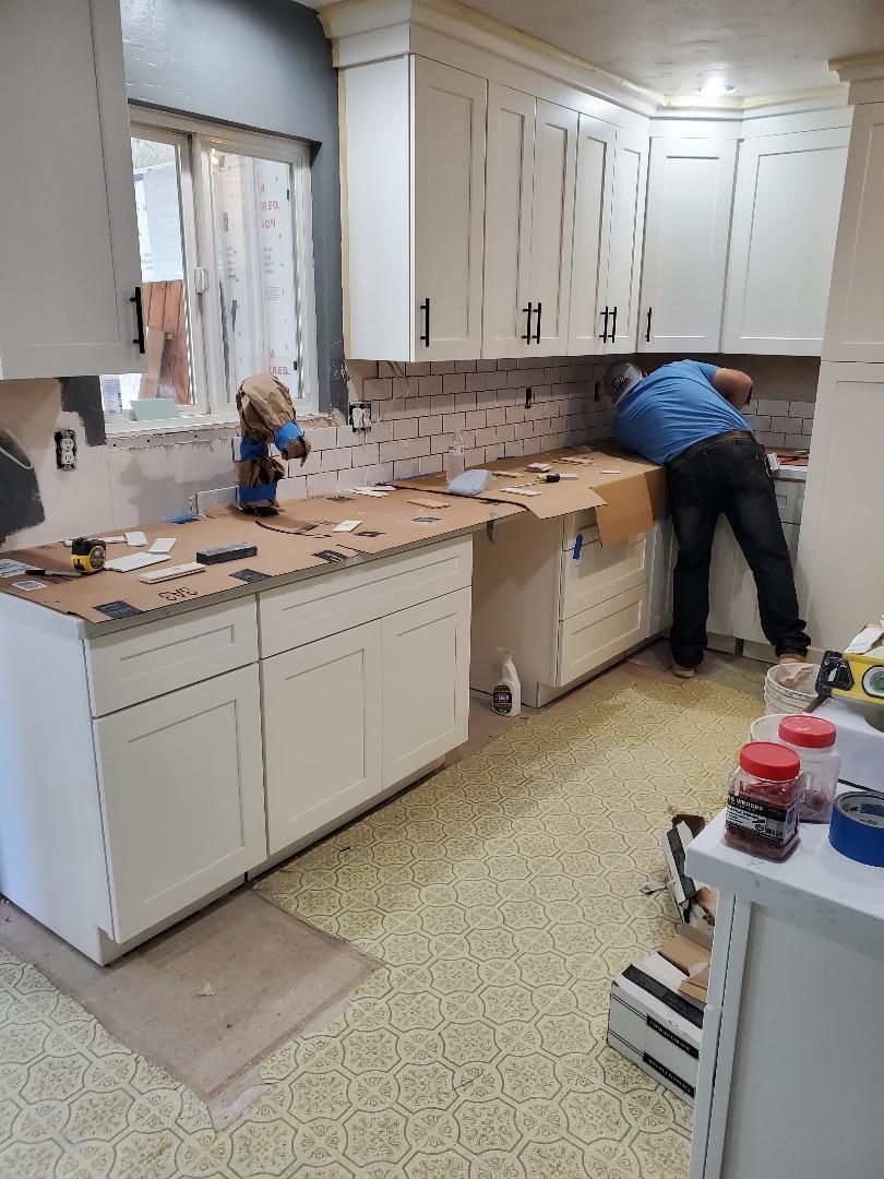 JJR Construction -  A person installing backsplash in a kitchen. White cabinets, beige flooring, and tools are visible.