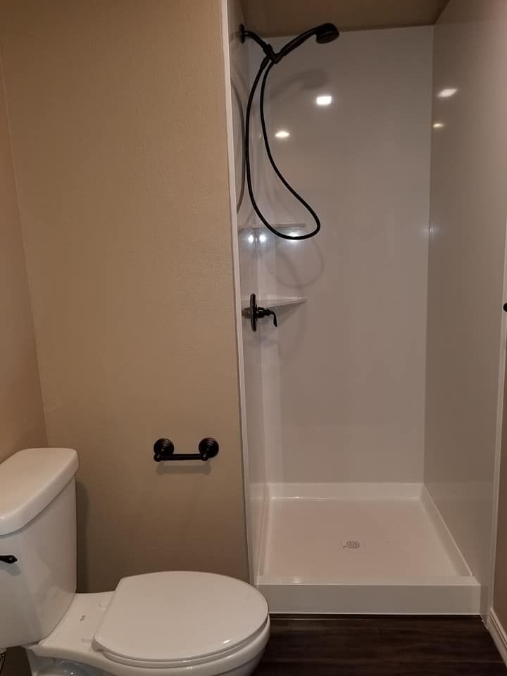 JJR Construction - Bathroom with white shower stall, toilet, and black fixtures. Beige walls and dark wood floor.