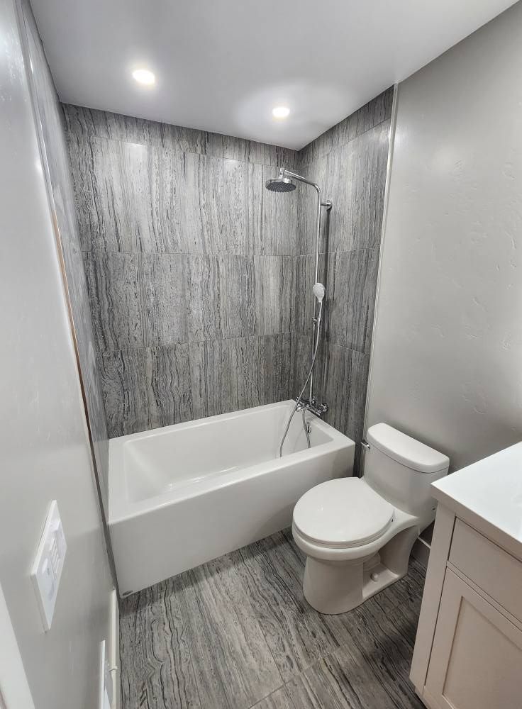JJR Construction -  Bathroom with white tub, toilet, and gray wood-look wall and floor panels.