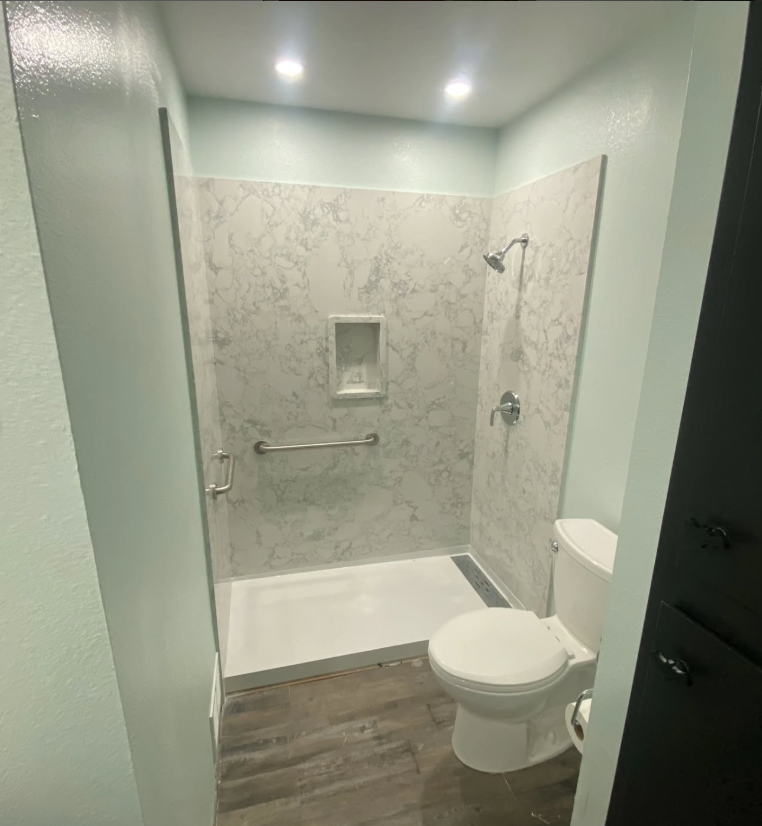 JJR Construction - Bathroom with shower, marble-look walls, grab bar, and toilet. Light green walls, gray floor, and recessed lighting.